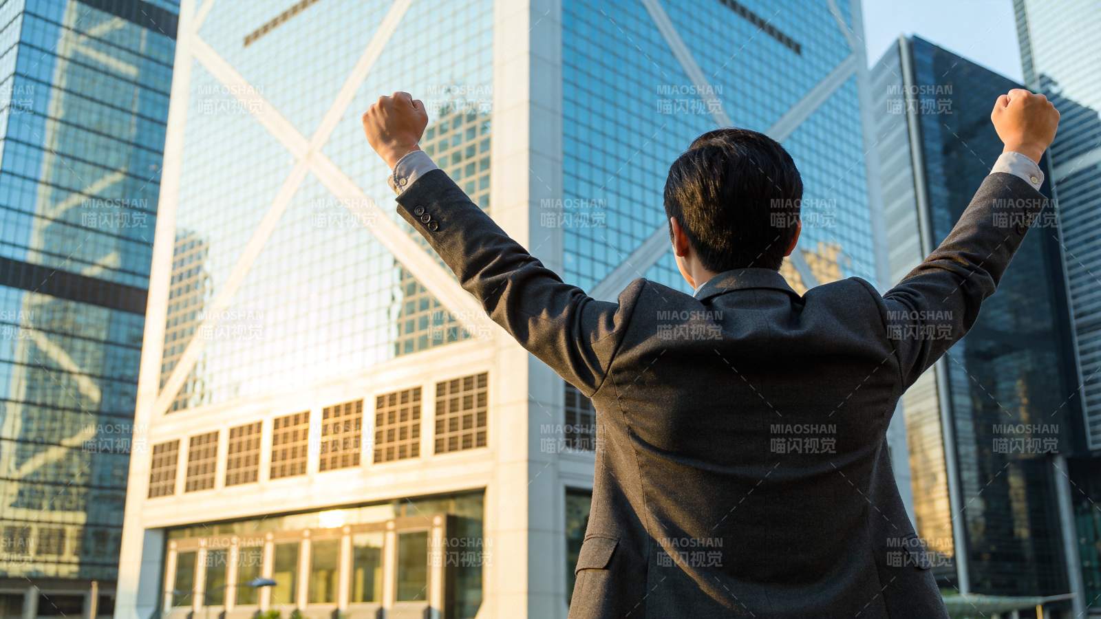 西装男子背对镜头双臂高举于摩天大楼前图片