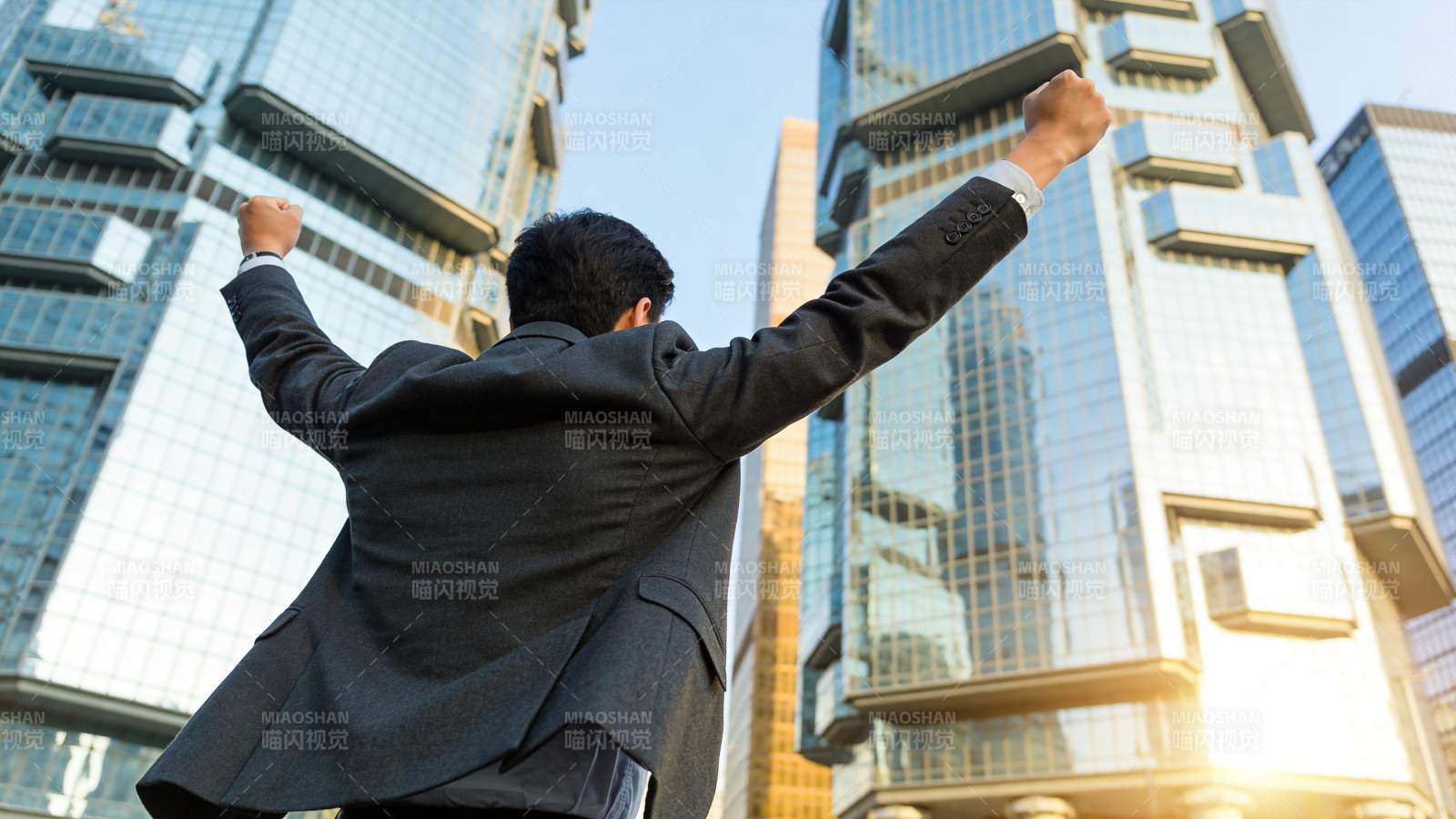 西装男子背对镜头双臂高举于摩天大楼前图片