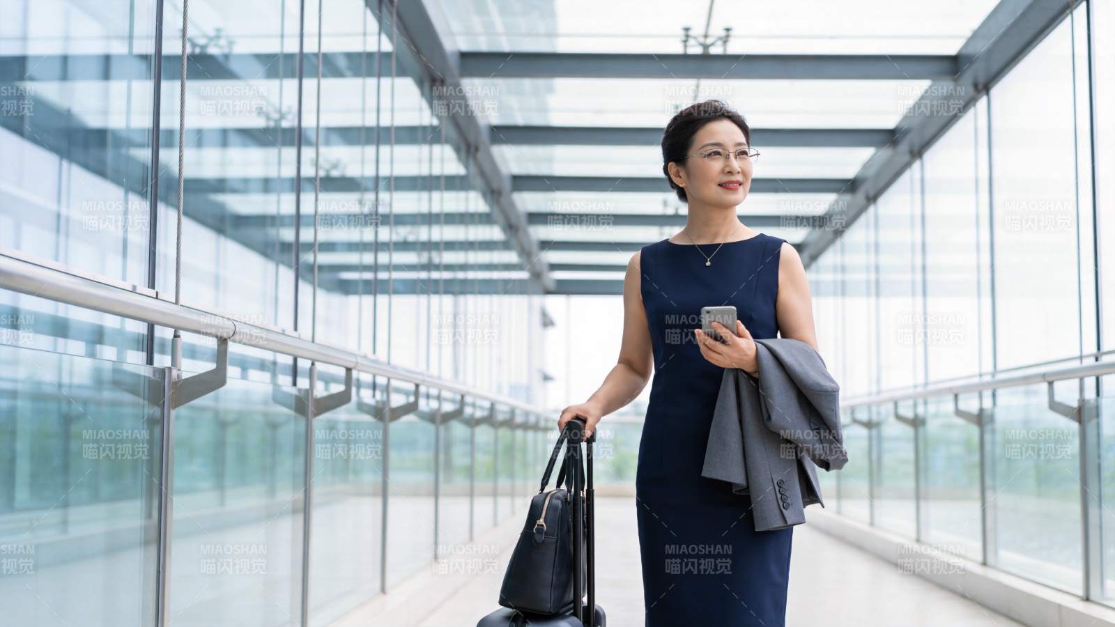 穿深蓝连衣裙女子手提行李箱行走于玻璃廊桥图片