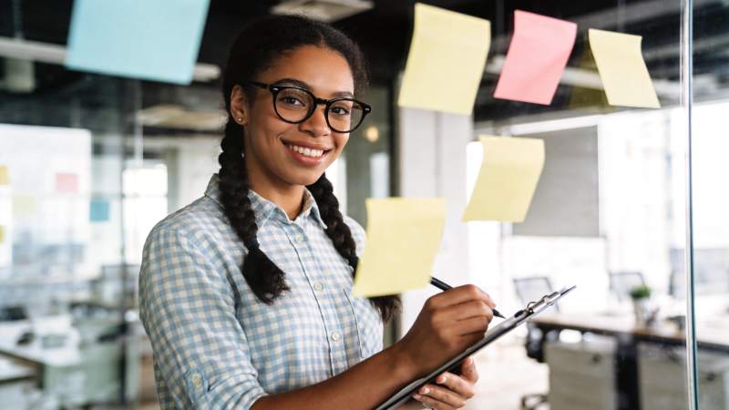 戴眼镜女性手持剪贴板站在贴满便签的玻璃前图片
